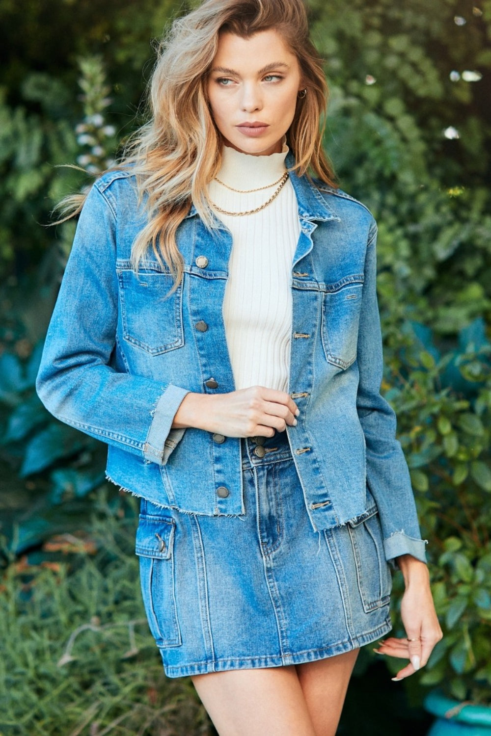 Trendy denim jacket with a raw hem and button-up design, worn by a stylish young woman with long wavy blonde hair in an outdoor natural setting.