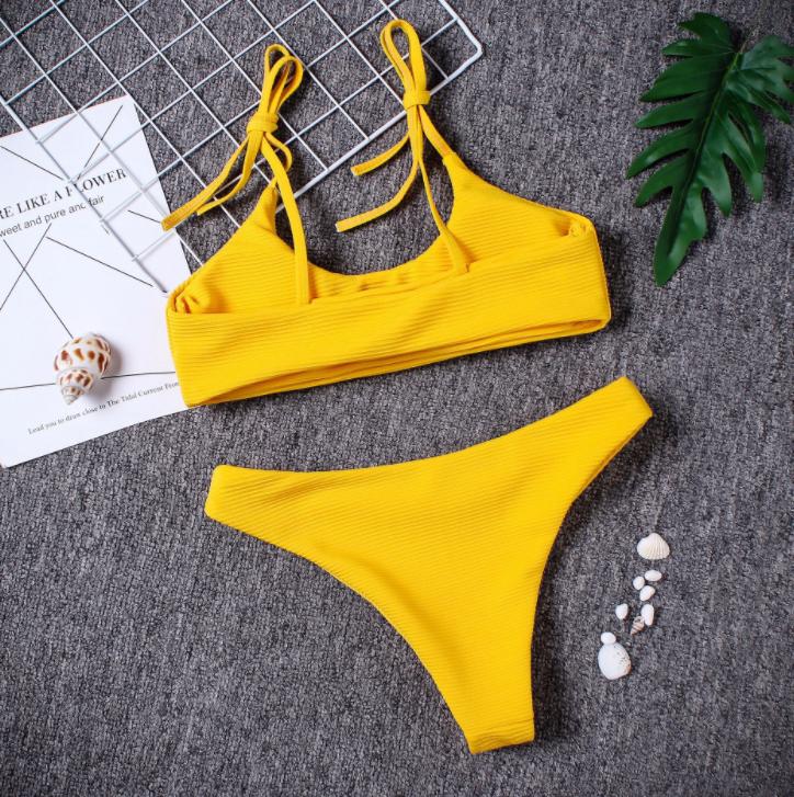 Vibrant yellow women's swimsuit with adjustable straps and a classic bikini bottom design, resting on a textured gray background with green leaves and seashells.