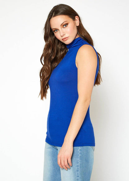 Sleeveless blue turtleneck top, fitted style, worn by young woman with long wavy brown hair posing against a plain white background.