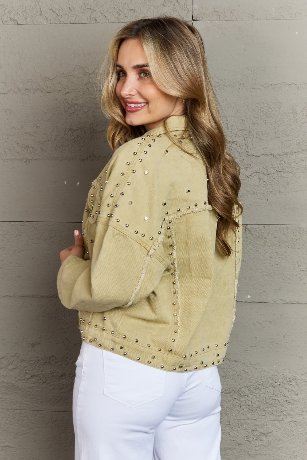 Stylish studded denim jacket with frayed trim, showcased by a smiling female model against a textured wall.