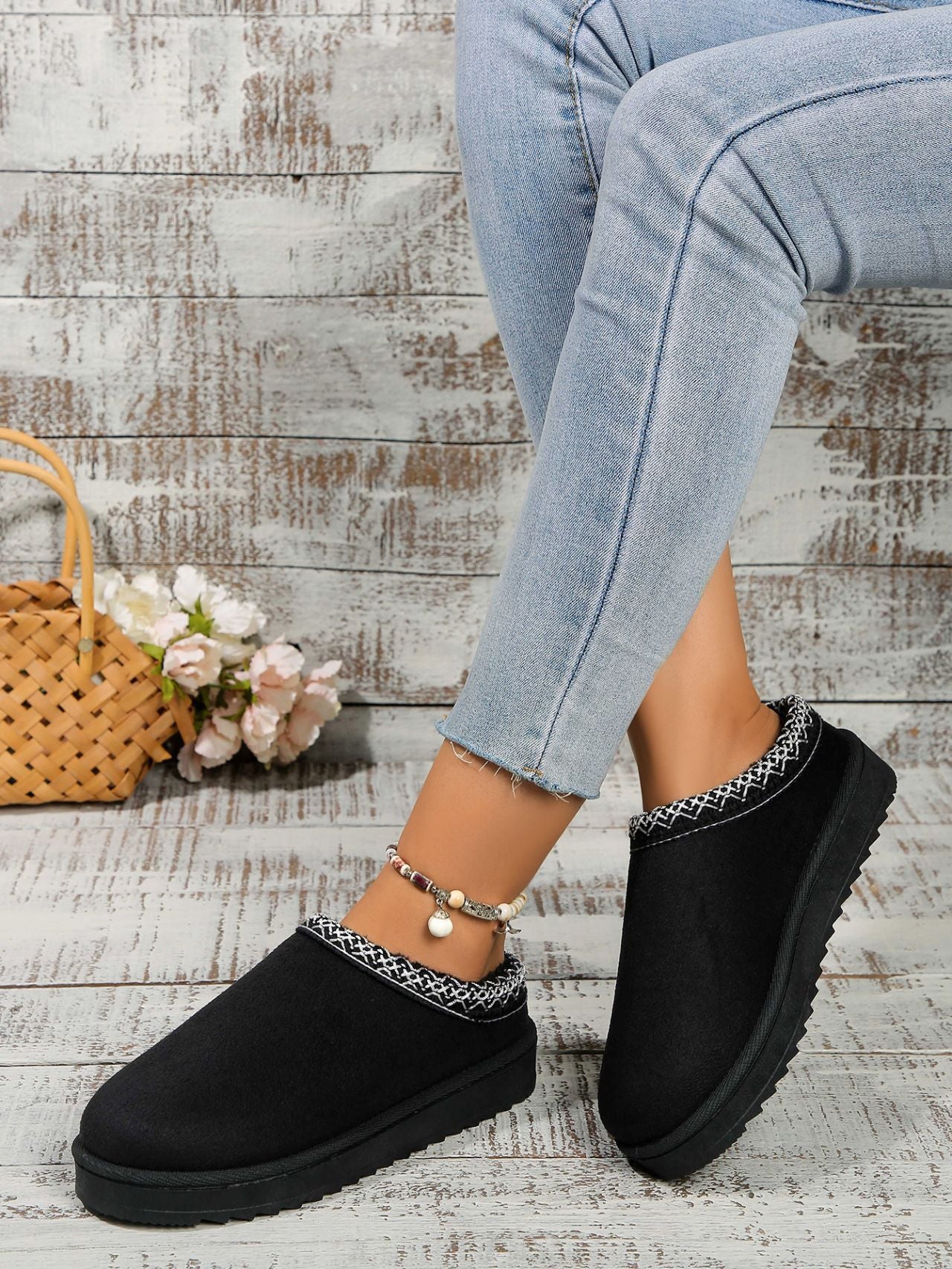 Round Toe Platform Slippers
Cozy black plush slippers with textured trim, resting on a rustic wood-paneled background adorned with a wicker basket and flowers.