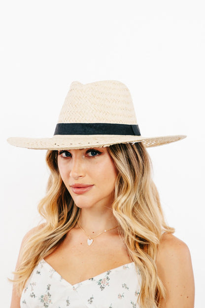 Fashionable woven straw sun hat with dark trim, worn by a young woman with long blonde hair against a plain white background.