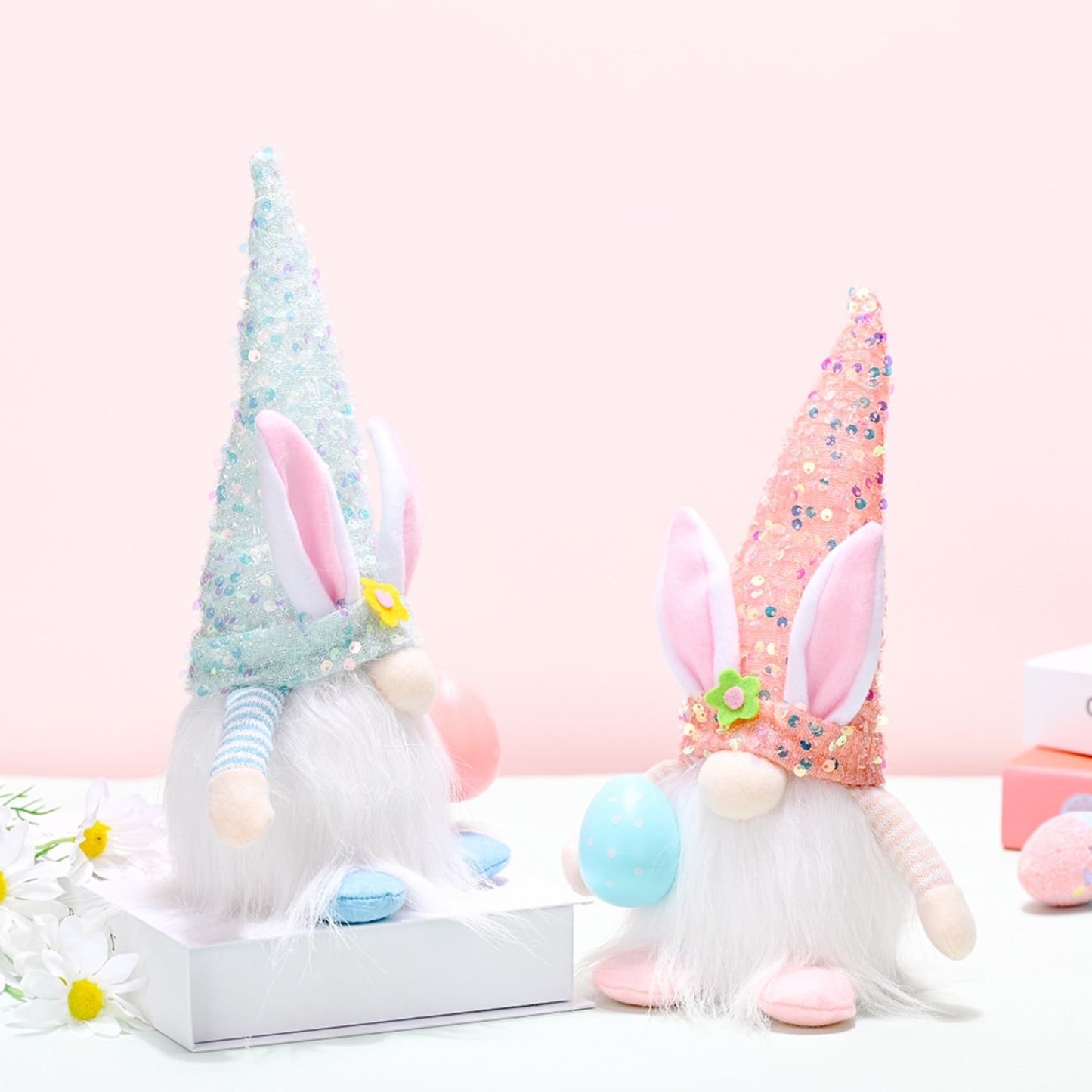 Whimsical Easter Gnome Figurines - Festive sequin hats and soft white fur bodies of two decorative Easter-themed gnome statues, one in soft pastel blue and the other in a bright multicolored pattern, sitting on a reflective white surface with flowers in the background.