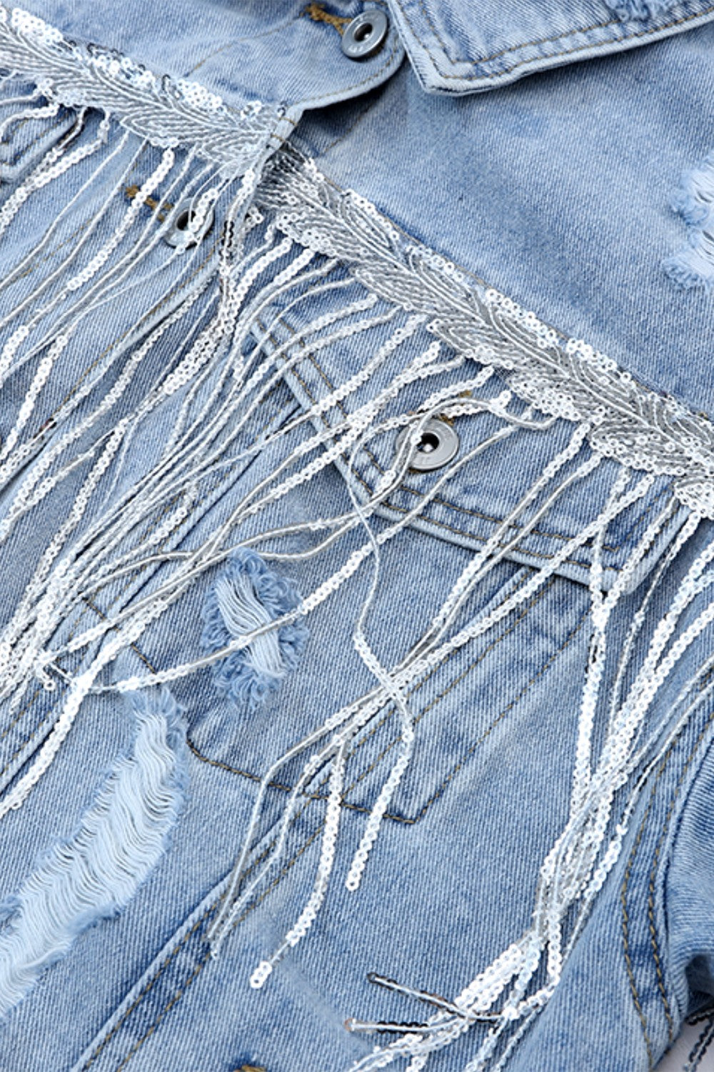 Distressed denim jacket with sequin fringe trim. The image shows the front of a light blue denim jacket with intricate sequin detailing and fringes along the collar and shoulders, creating a stylish and textured look.