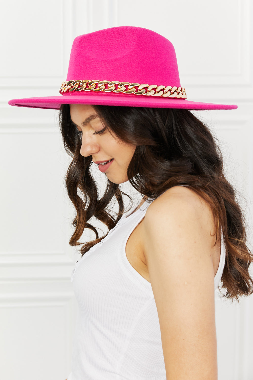 Elegant pink felt fedora hat with chain trim, worn by a model with long, wavy dark hair against a white background.