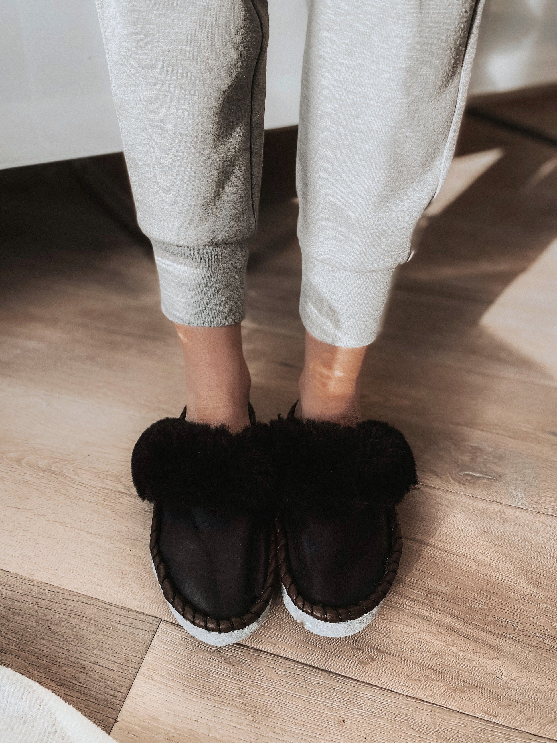 Cozy black faux fur slip-on slippers with white soles, worn over gray sweatpants on a wooden floor