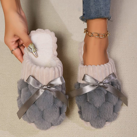 Soft white and gray faux fur slippers with ribbon bows, worn by a person with jeans and jewelry visible in the image.