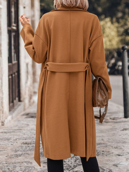 Elegant camel-colored long coat with tie waist, shown on a woman walking down a city street.