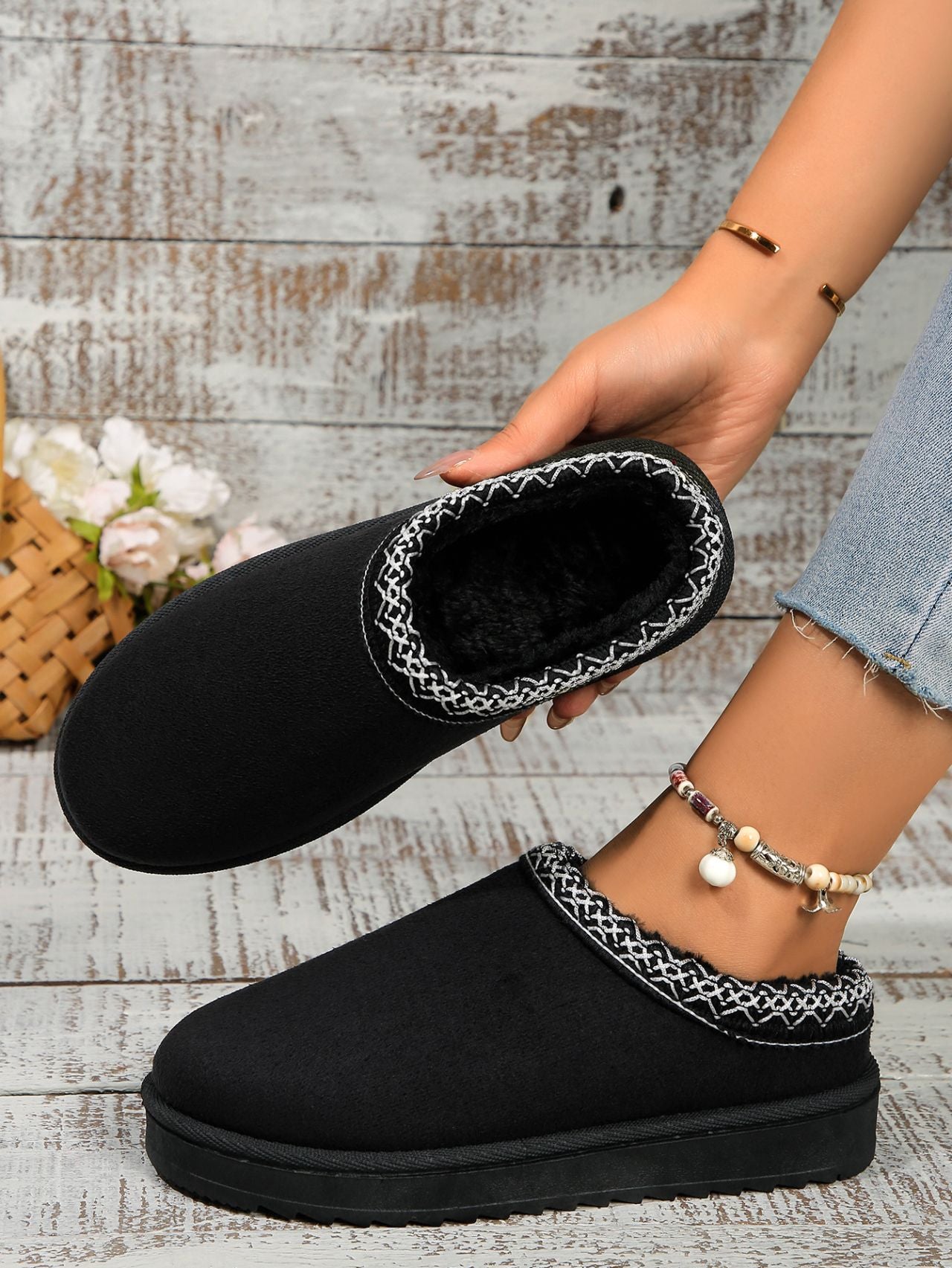 Cozy black velvet platform slippers with contrasting white stitching, showcased on a hand against a rustic wooden background with flowers and a wicker basket.