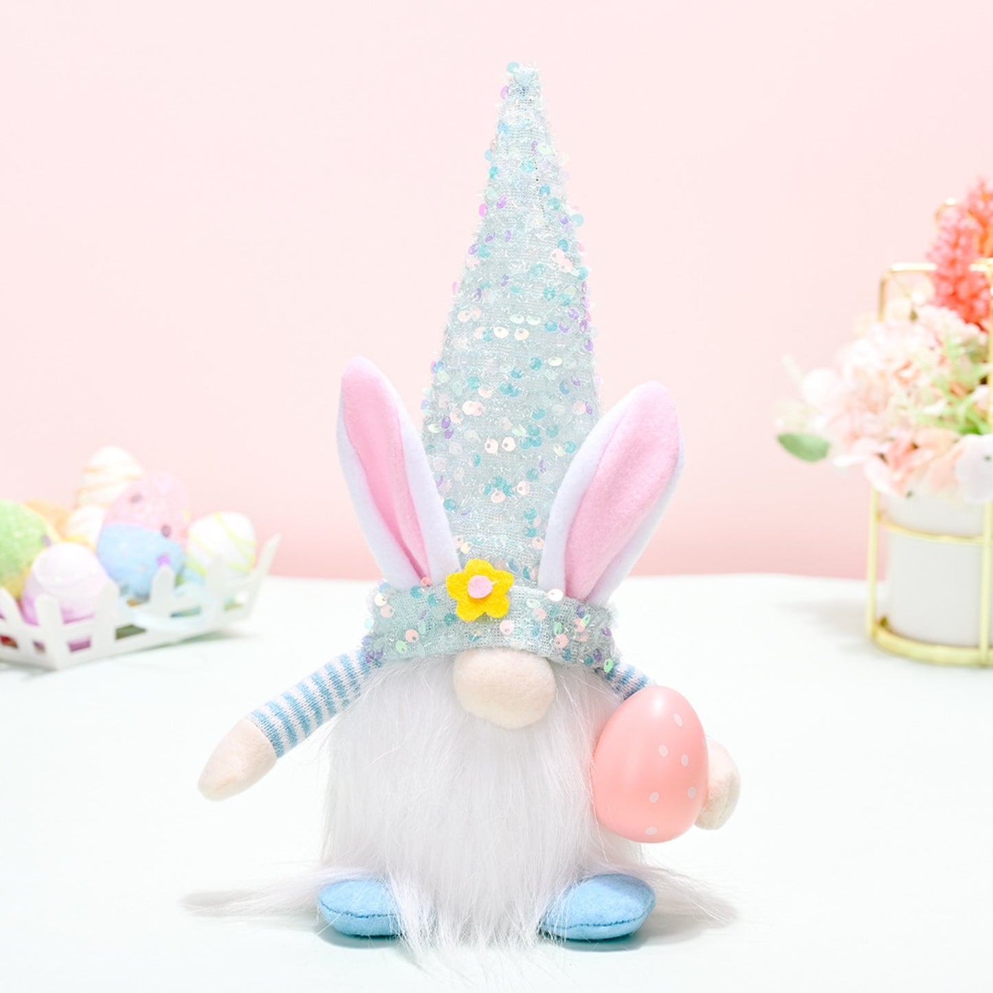 Glittering Easter Sequin Pointed Hat Faceless Gnome
A whimsical and festive Easter gnome figurine with a sparkling sequined hat, adorned with fluffy pink bunny ears. The gnome sits on a white surface, surrounded by pastel-colored Easter eggs and a vibrant floral arrangement, creating an endearing and cheerful spring-themed display.