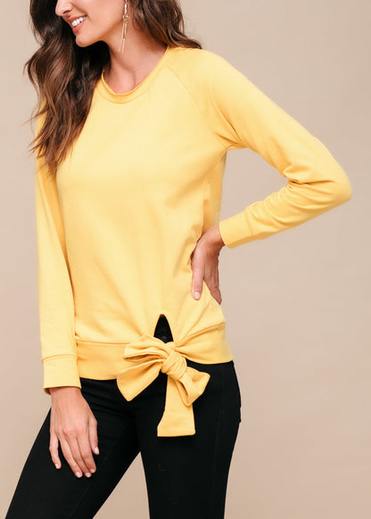 Yellow women's crew neck tie knot sweatshirt with long sleeves, worn by a smiling young woman with long brown hair against a plain background.