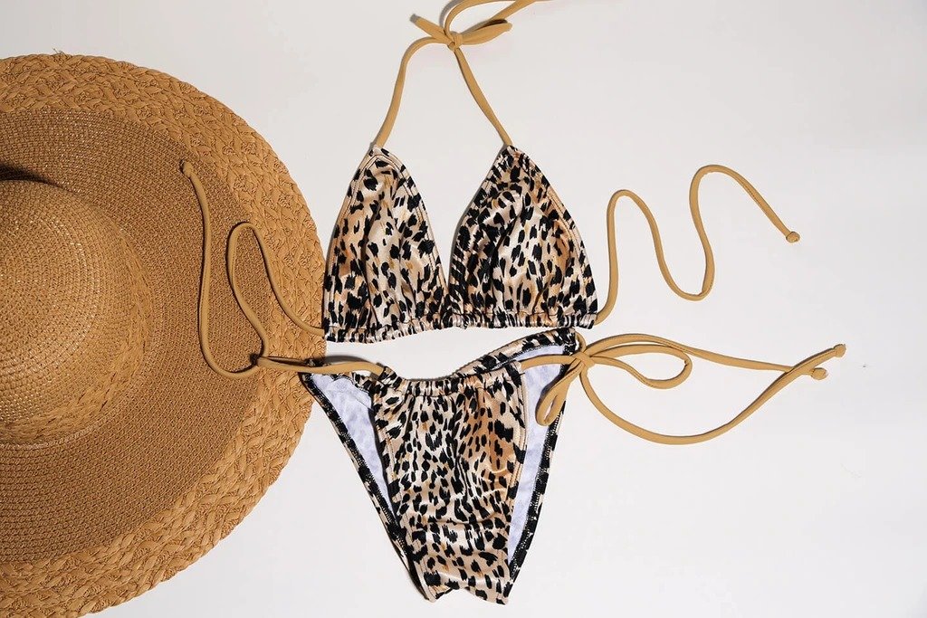 Leopard-print bikini with golden accents, straw hat in serene setting.