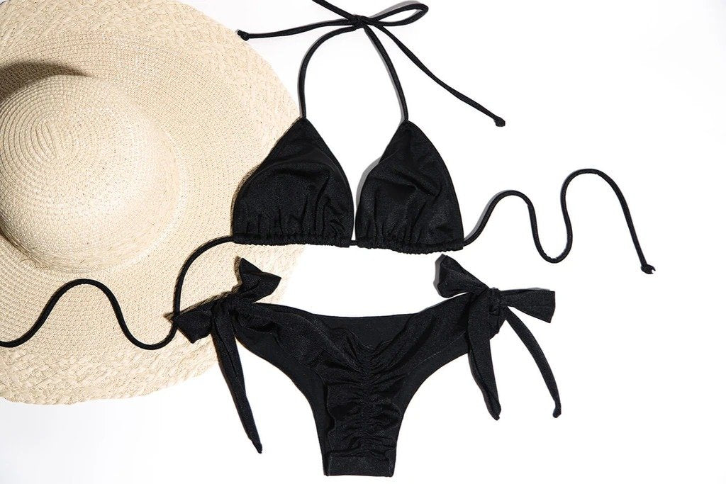 Stylish black bikini with string ties, accompanied by a chic wide-brimmed sun hat, captured against a plain background.