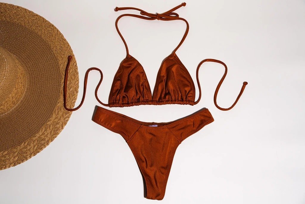 Rust-colored bikini with plunging neckline, halter straps, and high-cut bottoms on white background with straw hat.