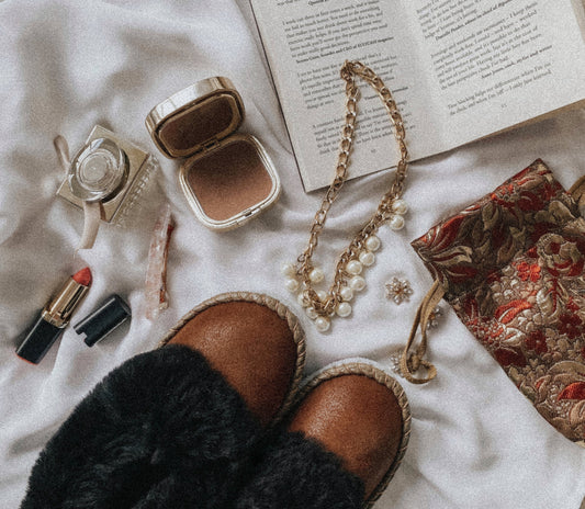 Cozy brown sheepskin slippers, makeup compact, lipstick, and pearl necklace arranged on a soft white surface with a book in the background.