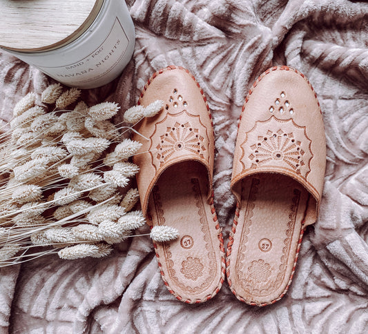 Detailed description of the product image:

Rustic Embroidered Leather Slippers on Cozy Blanket

The image shows a pair of closed-toe leather slippers with delicate embroidered designs on a soft, textured blanket. The slippers have a warm, earthy tone and a decorative pattern that creates a rustic, handmade aesthetic. They are surrounded by natural elements like dried flowers, adding to the cozy, artisanal feel of the scene.