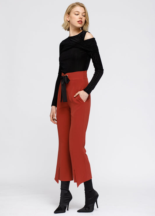 Elegant woman's high-waist front slit trousers in a rich orange-red color, paired with a black cold-shoulder sweater, standing on white background.