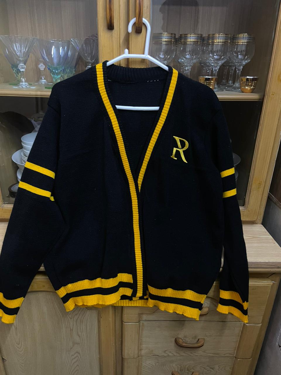 Stylish varsity-inspired women's cardigan in black with yellow trim, featuring the letter "R" for a bold, sporty look. Displayed on a hanger against a backdrop of shelves holding glassware.