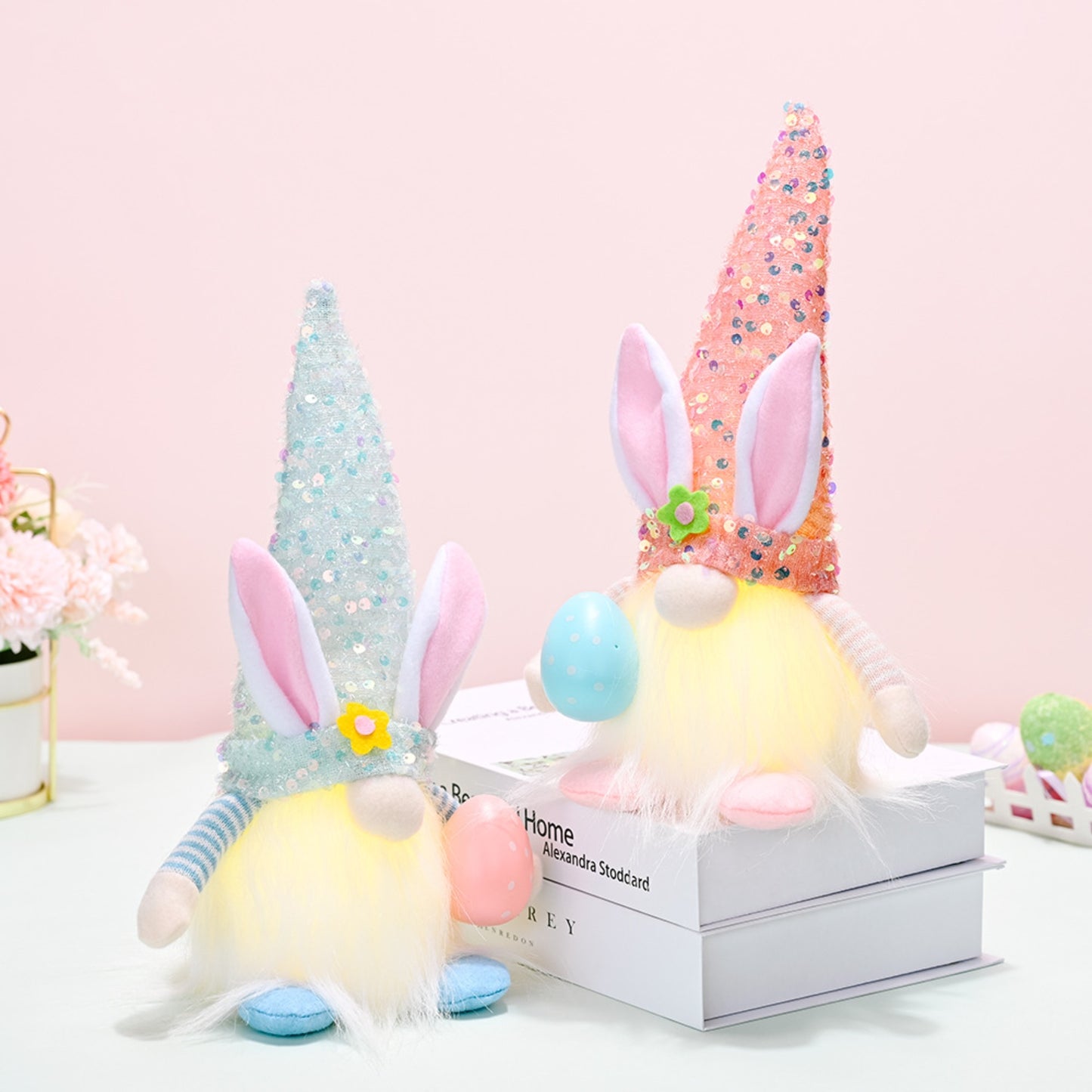 Colorful Easter gnomes with rabbit ears and sequin hats sitting on a white box surrounded by decorative flowers and pastel eggs