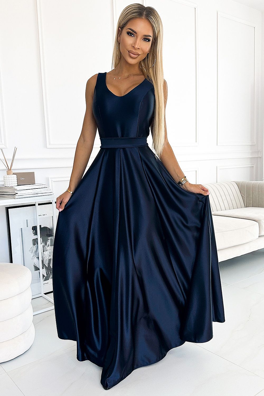 Elegant navy blue satin ball gown dress with a V-neckline and flowing skirt, worn by a smiling young woman against a bright, minimalist background.