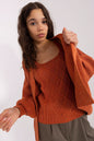 Cozy knit sweater in warm, rusty orange tones with intricate diamond pattern, modeled by a young woman with long, dark curly hair against a plain white background.