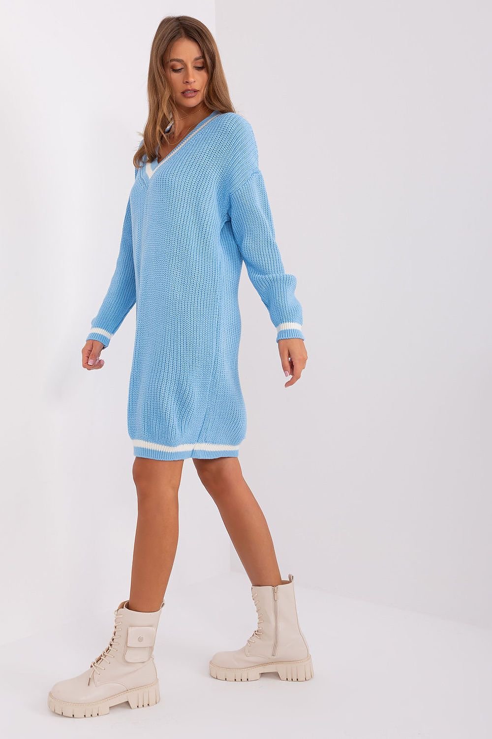 Soft blue knitted dress with long sleeves, worn by a young woman with long brown hair posing against a plain white background. The dress features a classic ribbed pattern and an oversized, relaxed silhouette. The model is also wearing chunky white boots, completing the casual yet stylish look.