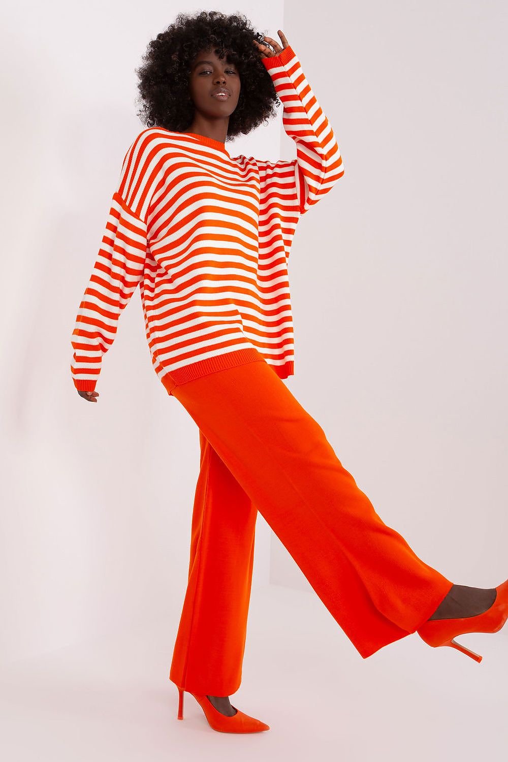 Striped knitted sweater, wide-leg orange pants, and red heels featured in image of a woman with curly hairstyle posing.