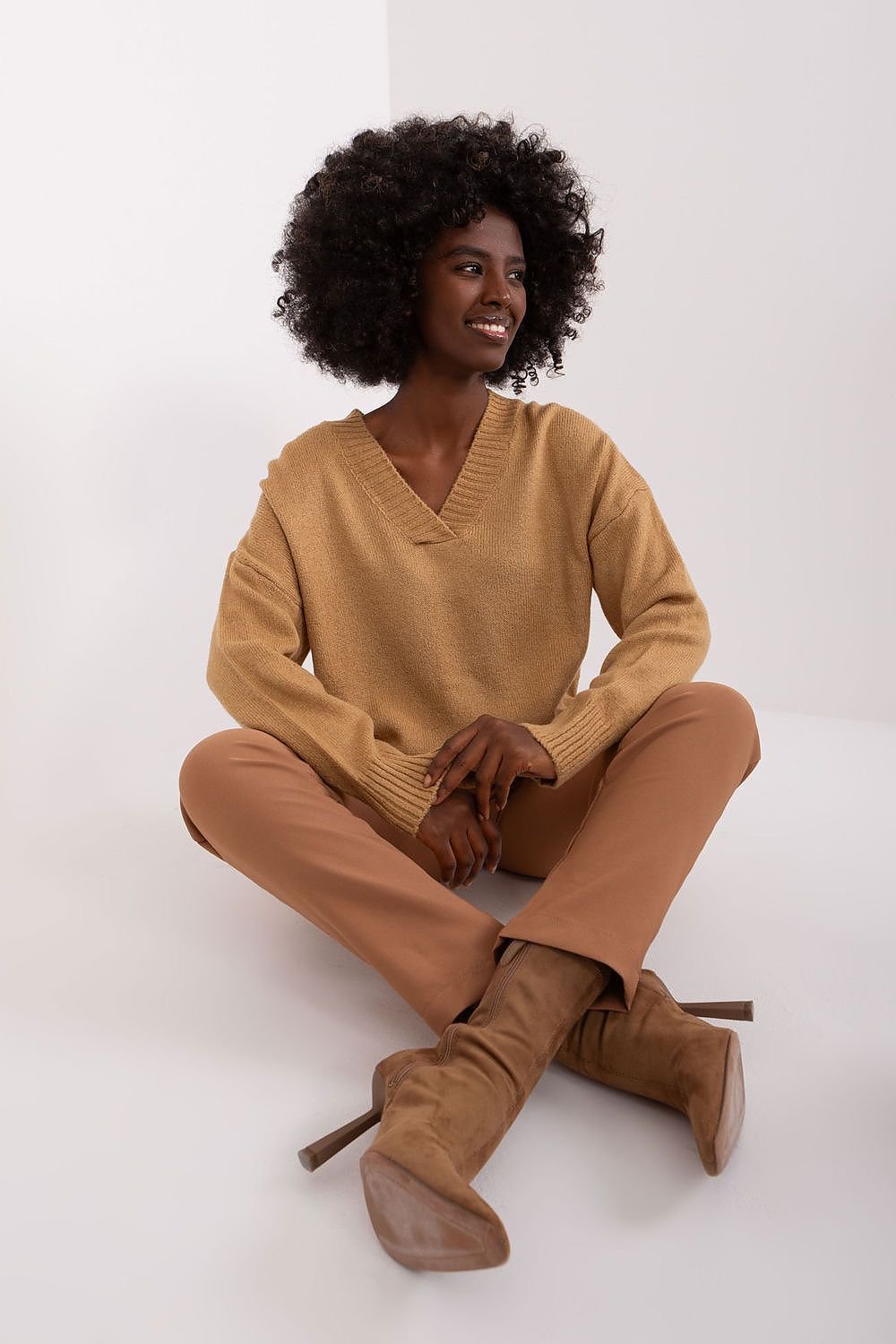 Cozy brown jumper with v-neck style, worn by a smiling woman with curly dark hair sitting on white background.