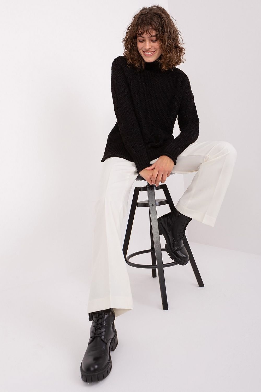 Curly-haired woman in cozy black turtleneck sweater, white pants, and heavy boots sitting on a stool against a plain background.