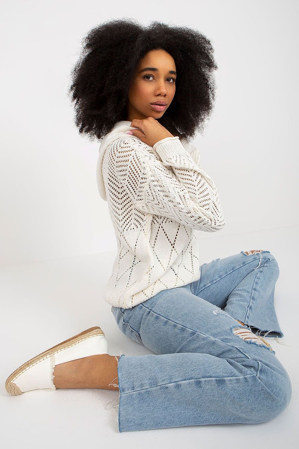 Elegant woman with curly dark hair wearing a cozy white knit sweater and blue jeans.