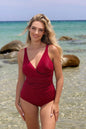 Elegant red one-piece swimsuit with flattering ruched details, worn by a smiling young woman with windblown blonde hair on a tropical beach.