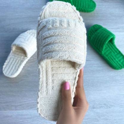 Soft, plush white and silver furry slippers held in woman's hand on wooden surface with green slippers in background.