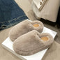 Soft, cozy beige fuzzy round toe flat slippers on a white surface, beside a pair of jeans, showcasing their comfortable and stylish design.