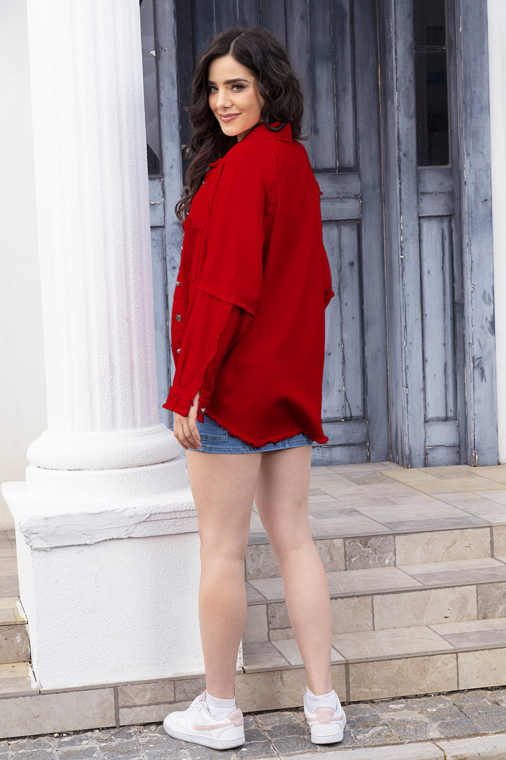 Bright red collared button-down jacket, relaxed denim shorts, and white sneakers on woman posing outdoors against weathered wooden wall.