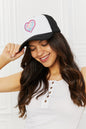 Stylish black and white trucker cap with a colorful heart design, worn by a smiling woman with long, wavy brown hair against a plain white background.