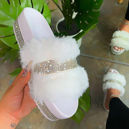 Fluffy white furry slippers with rhinestone-embellished straps, displayed on a hand against a background of lush green leaves.