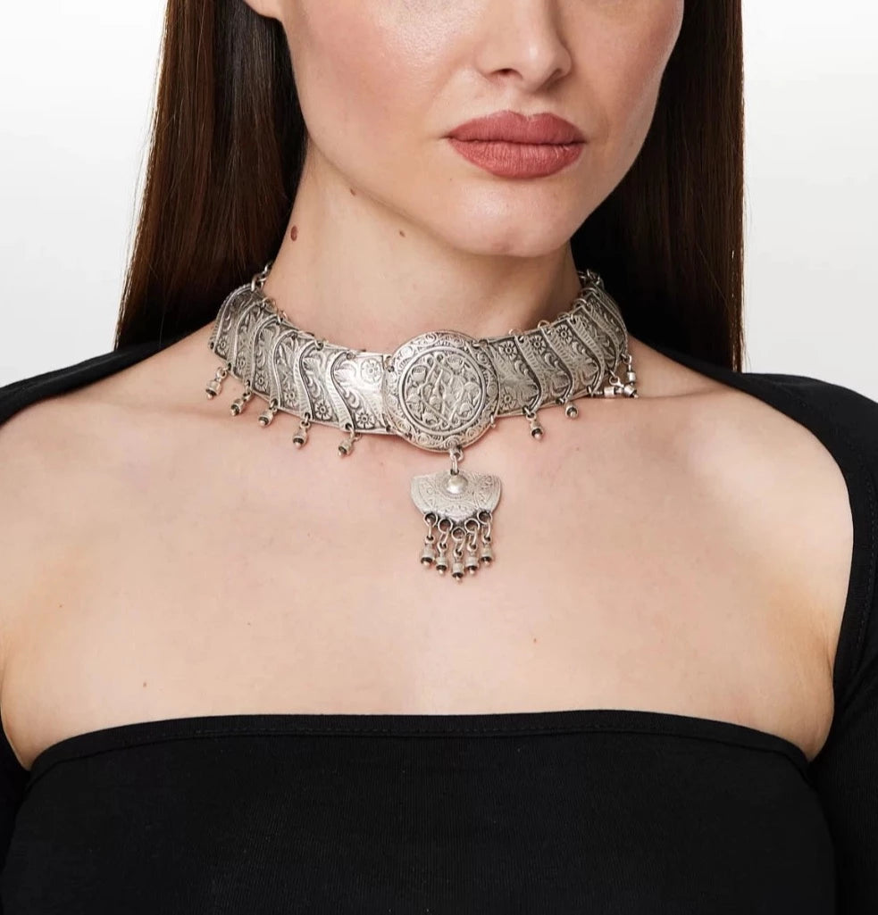 Ornate silver choker with intricate filigree designs and dangling charms, showcased on a female model with long dark hair and an off-the-shoulder black top.